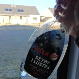 NS-CLEAN Kamila Wysocka - Butelka płynu do szyb 'Villa' w dłoni na tle okna i zabudowań. Widoczne krople na butelce sugerują świeże użycie. Słoneczny dzień.