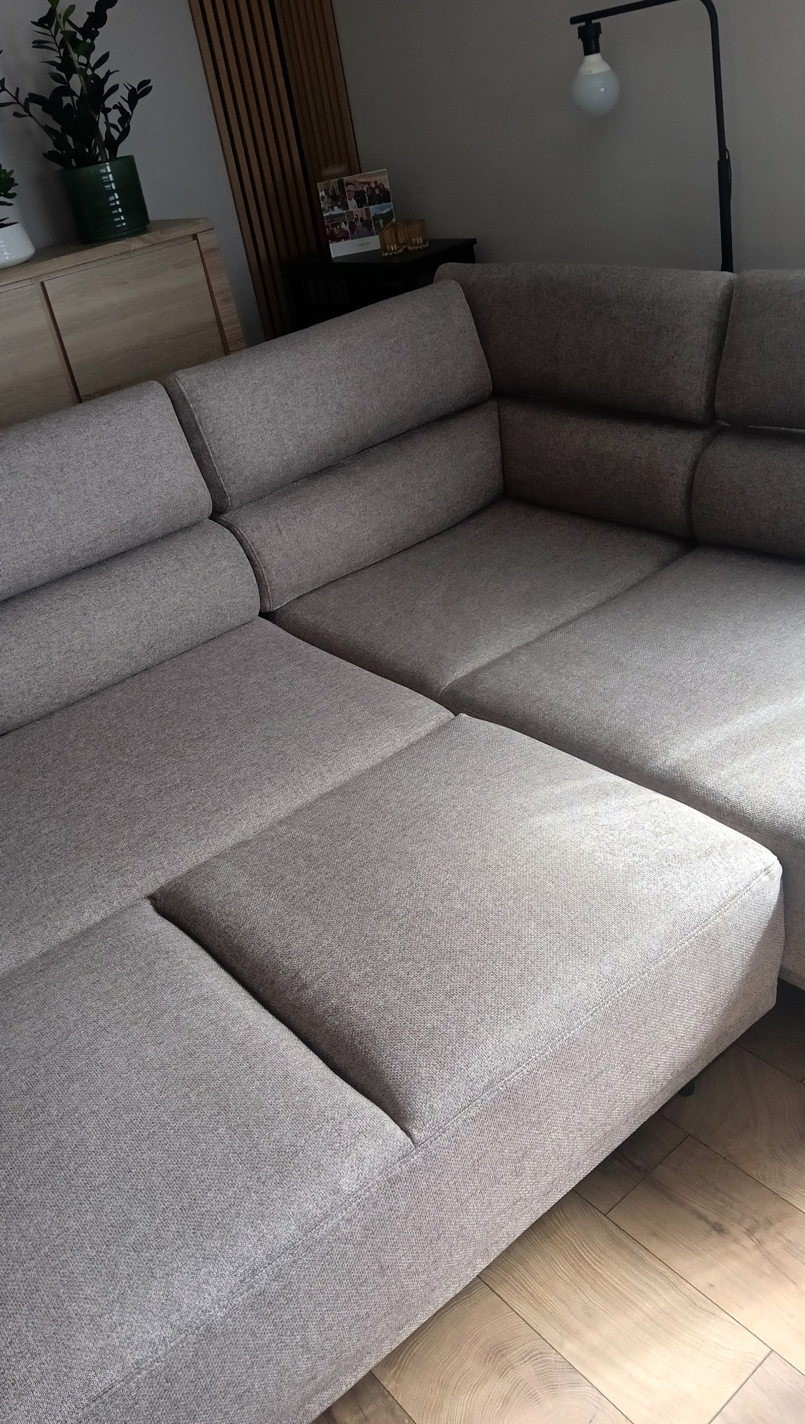 Jasnoszara, modułowa kanapa narożna z tkaniny o wyraźnym splocie, ustawiona w rogu pokoju z drewnianą podłogą i lampą w tle. Widoczne detale szwów i faktury materiału.