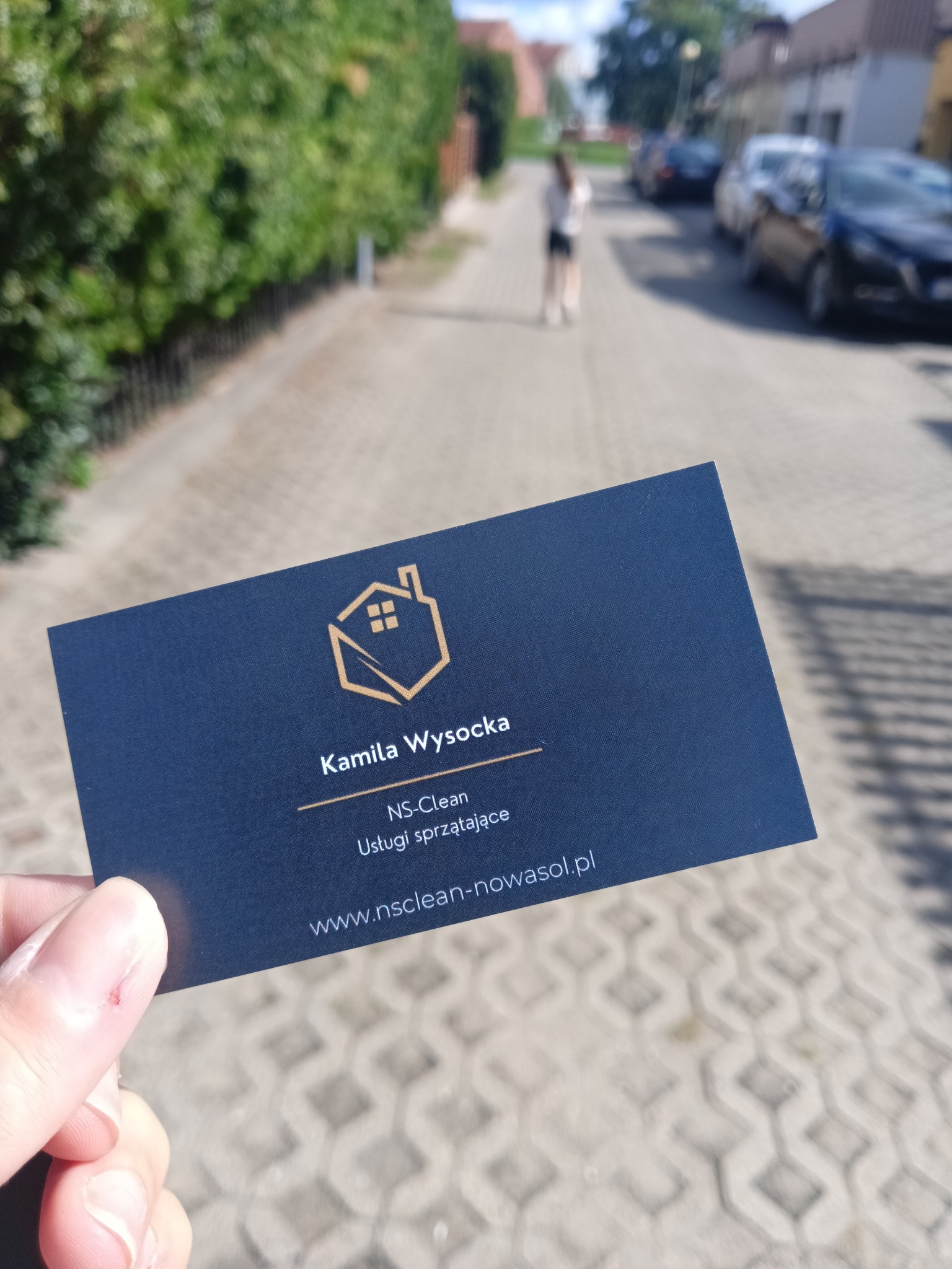 Granatowa wizytówka firmy sprzątającej NS-Clean Kamila Wysocka na tle ulicy z kostki, zieleni i oddalonych budynków.