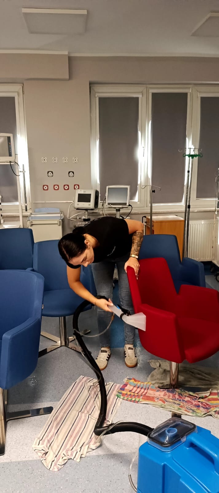 Pracownica z tatuażem pierze czerwone krzesło w pomieszczeniu z niebieskimi krzesłami i sprzętem medycznym. Widoczny niebieski odkurzacz piorący.