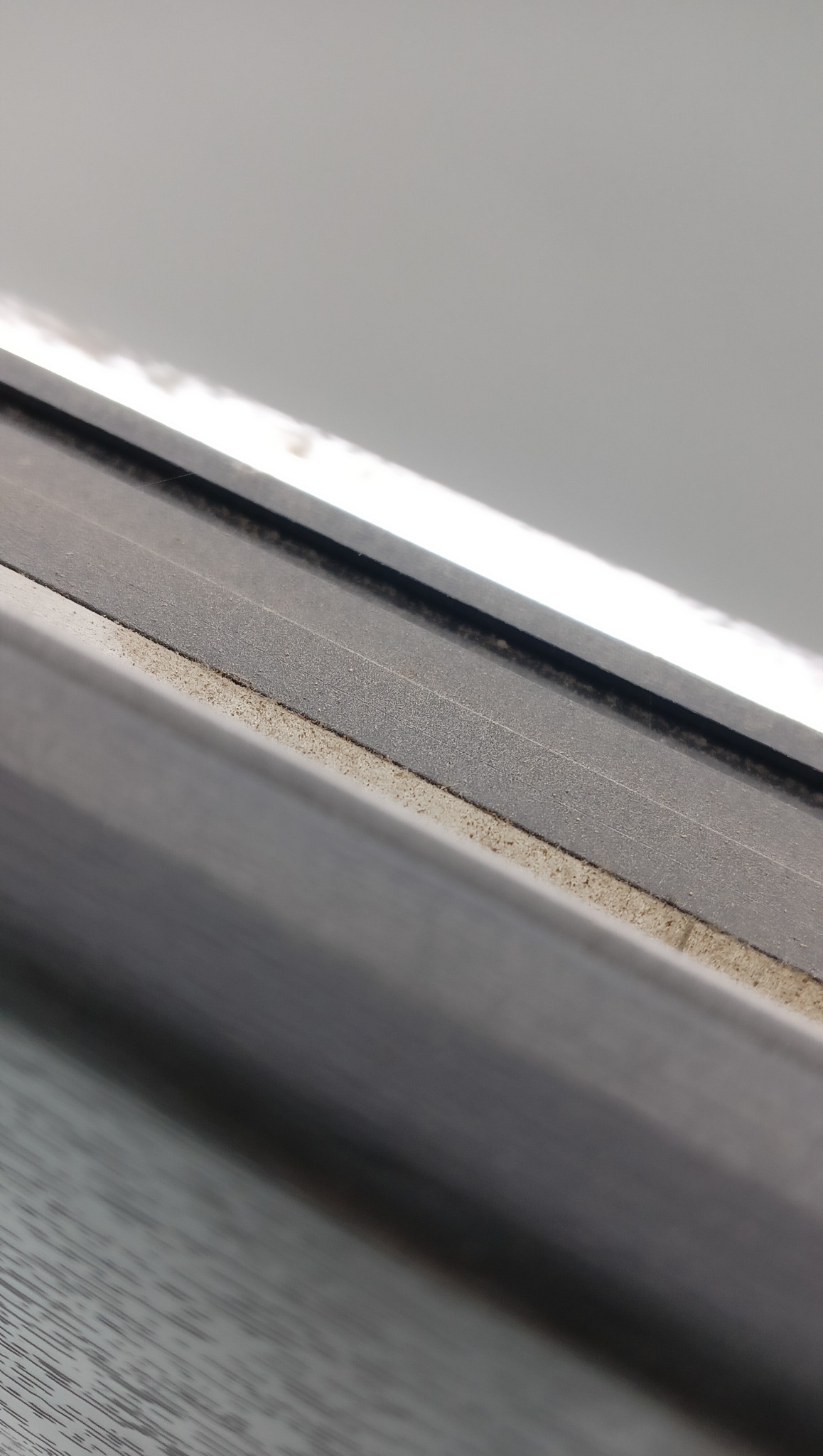 Brudna rama okienna z widocznymi drobinkami kurzu i zanieczyszczeń wzdłuż krawędzi. Tekstura ramy jest matowa, a światło pada z góry, podkreślając nierówności.