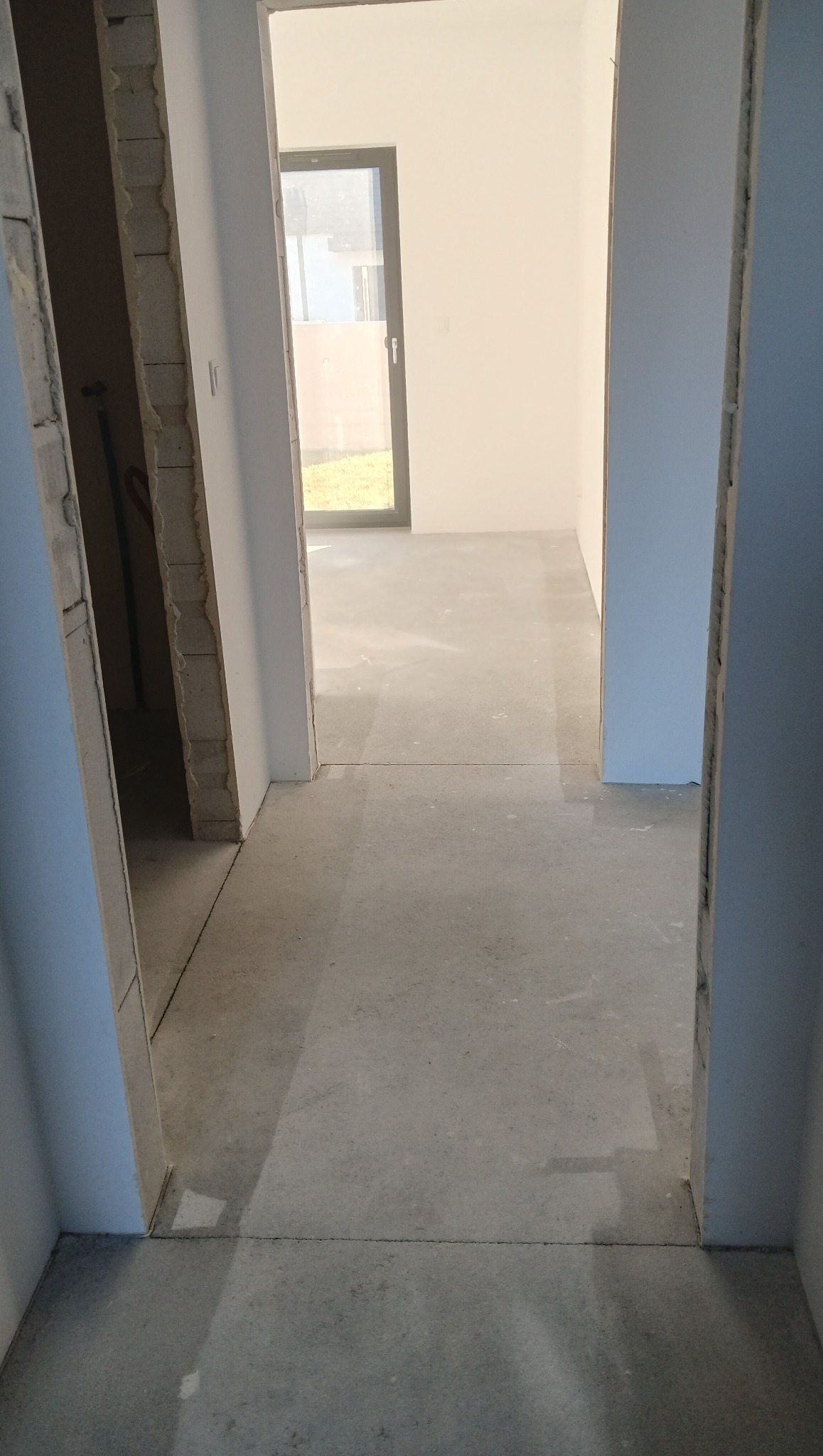 Surowy beton na podłodze w nowym mieszkaniu, widok przez dwa otwory drzwiowe. W tle okno z widokiem na zieleń. Ściany w stanie deweloperskim, gotowe do wykończenia.