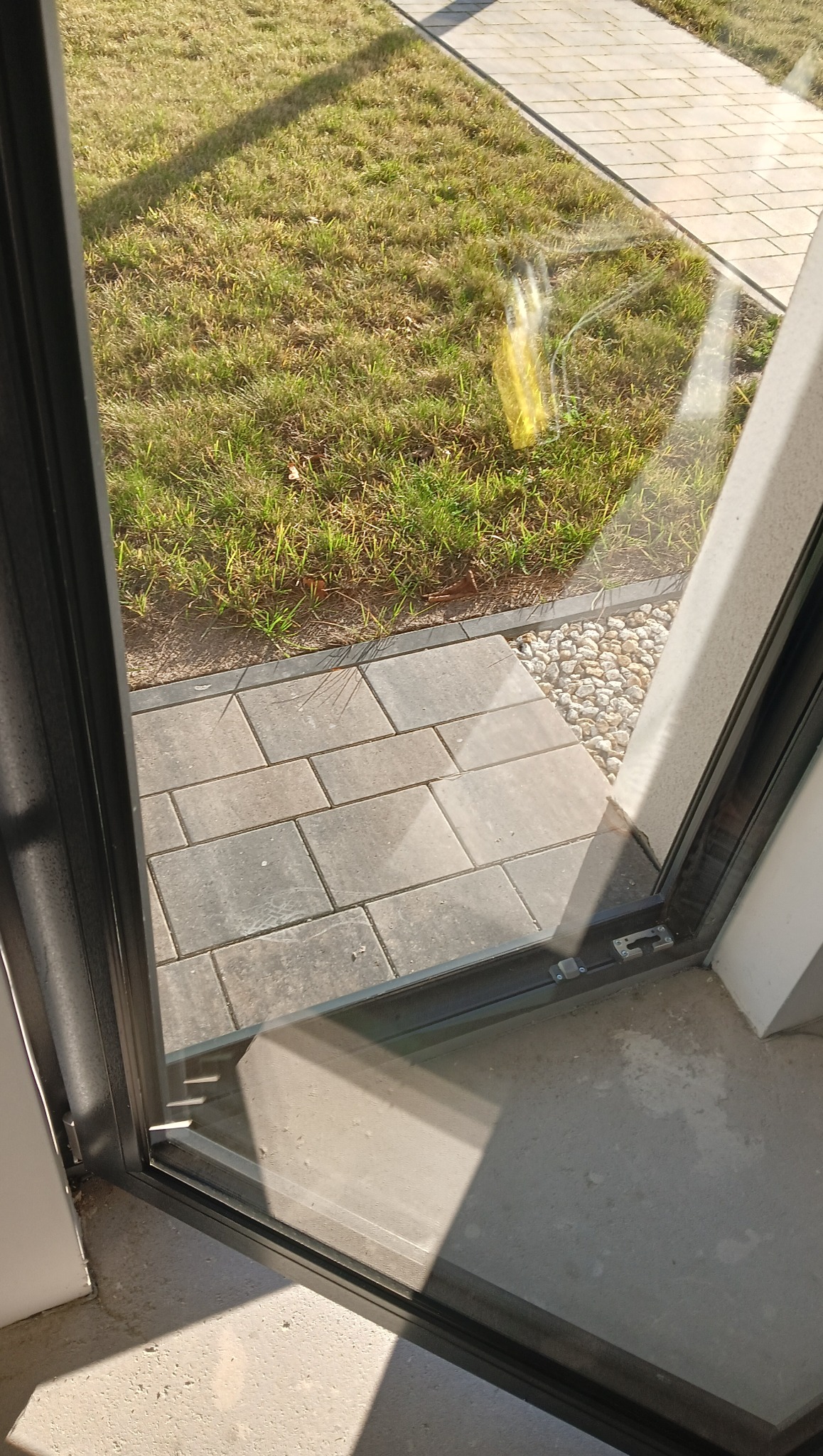 Widok przez otwarte, nowoczesne okno na trawnik i ścieżkę z kostki brukowej. Na szybie widoczne refleksy światła. Minimalistyczny design ramy okiennej.