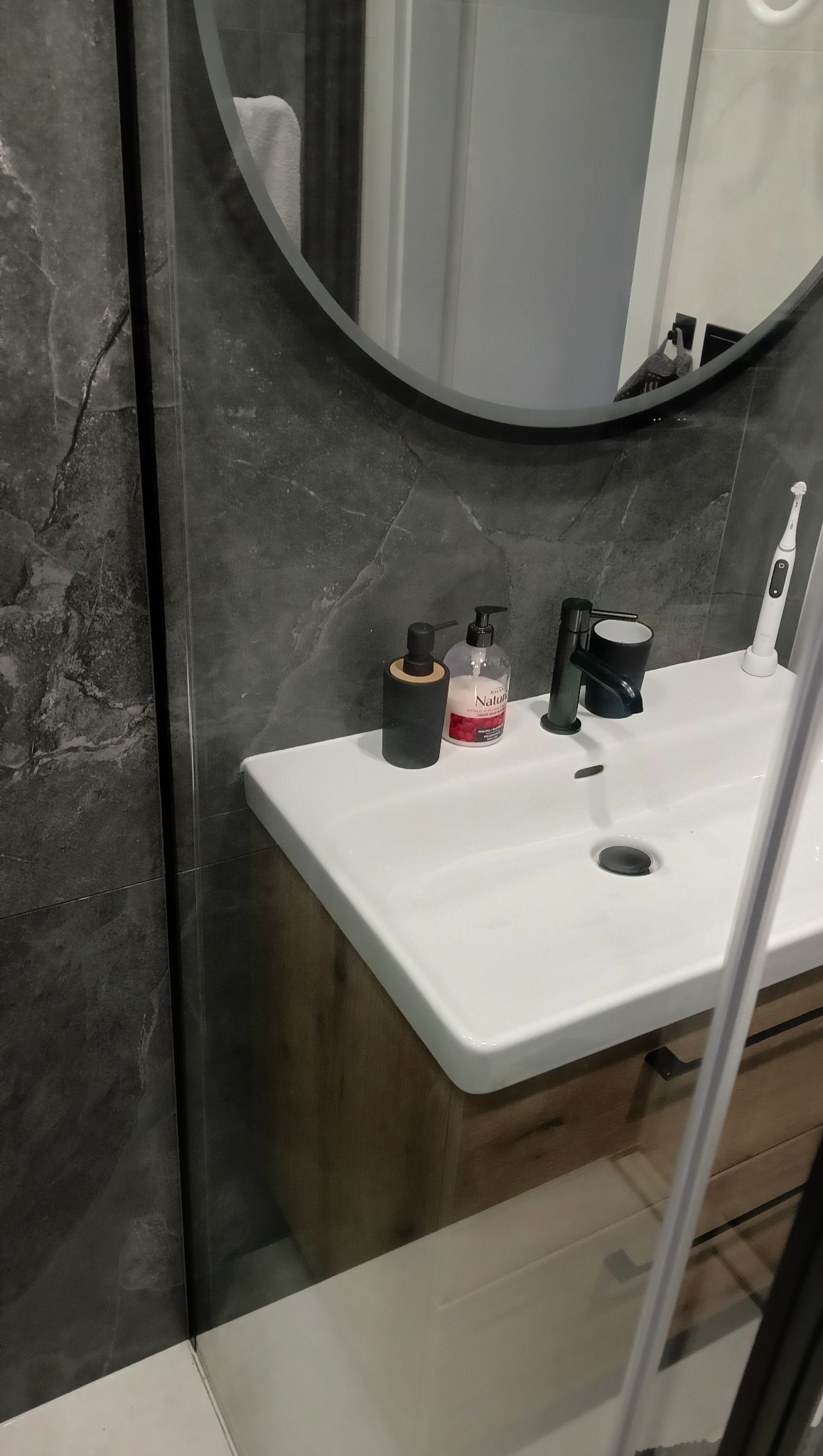 Nowoczesna łazienka z umywalką, czarną armaturą i dozownikami mydła na tle szarej ściany z efektem kamienia. Minimalistyczny design, czystość i porządek.