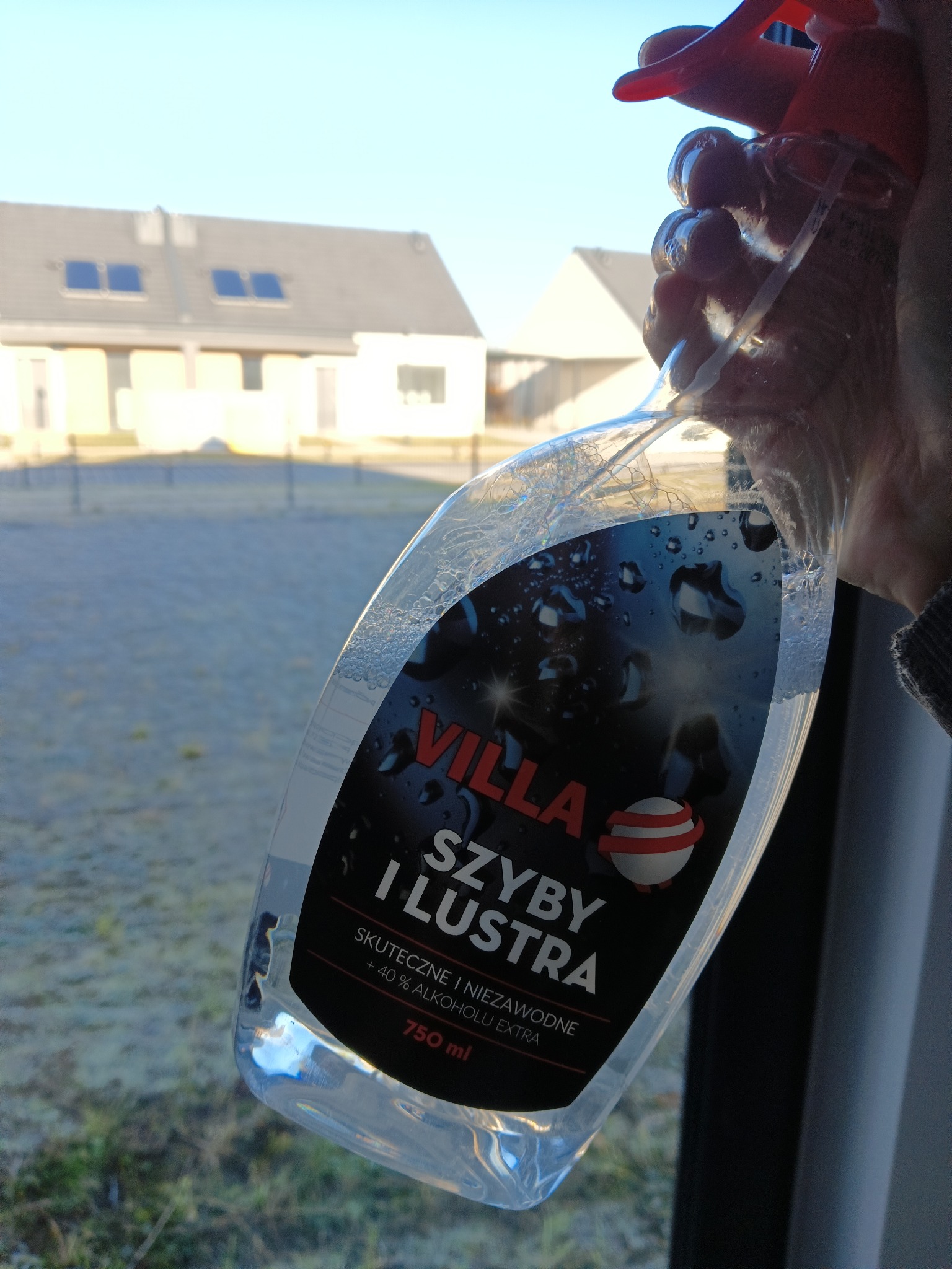 Butelka płynu do szyb 'Villa' w dłoni na tle okna i zabudowań. Widoczne krople na butelce sugerują świeże użycie. Słoneczny dzień.