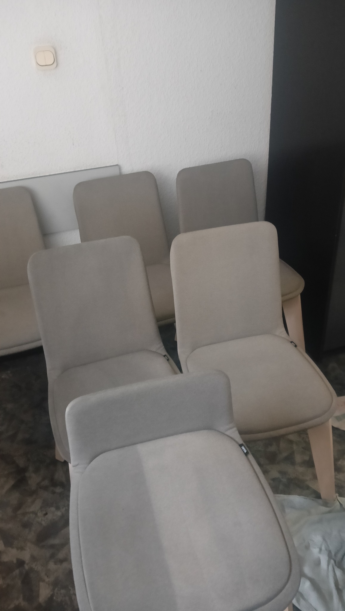 Jasnoszare krzesła tapicerowane z drewnianymi nogami ustawione w rzędzie, widoczne z bliska, z lekkim kątem z góry. Ściana w tle z włącznikiem.