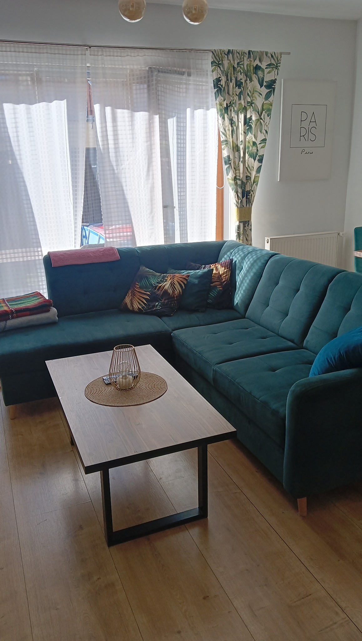 Szmaragdowa kanapa narożna z poduszkami w salonie, drewniany stolik kawowy z dekoracją, jasne zasłony w oknie, plakat 'Paris' na ścianie, parkiet na podłodze.