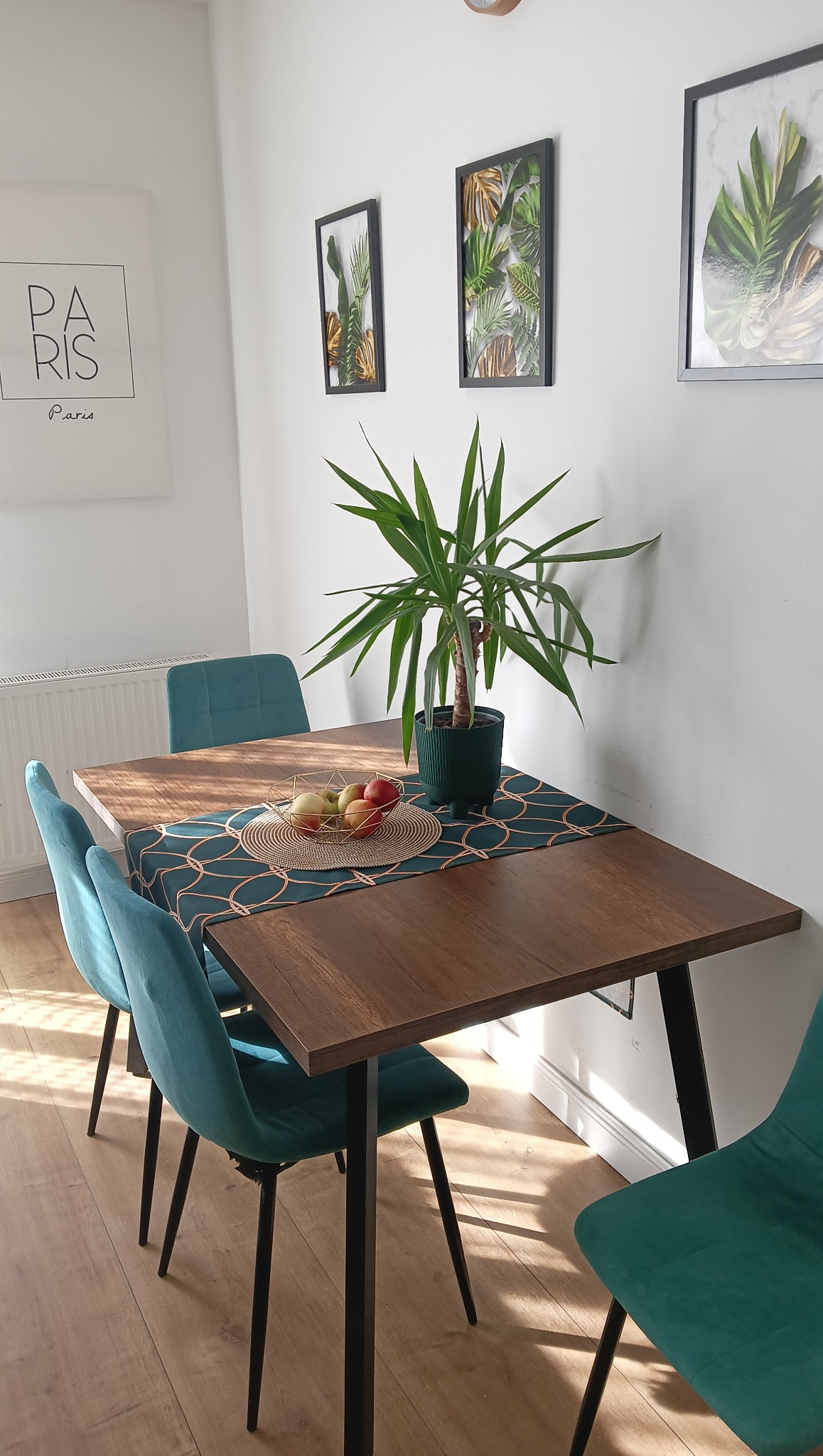 Jasne wnętrze jadalni: drewniany stół z turkusowymi krzesłami, dekoracyjny bieżnik, roślina w doniczce i grafiki na ścianie. Minimalistyczny design.