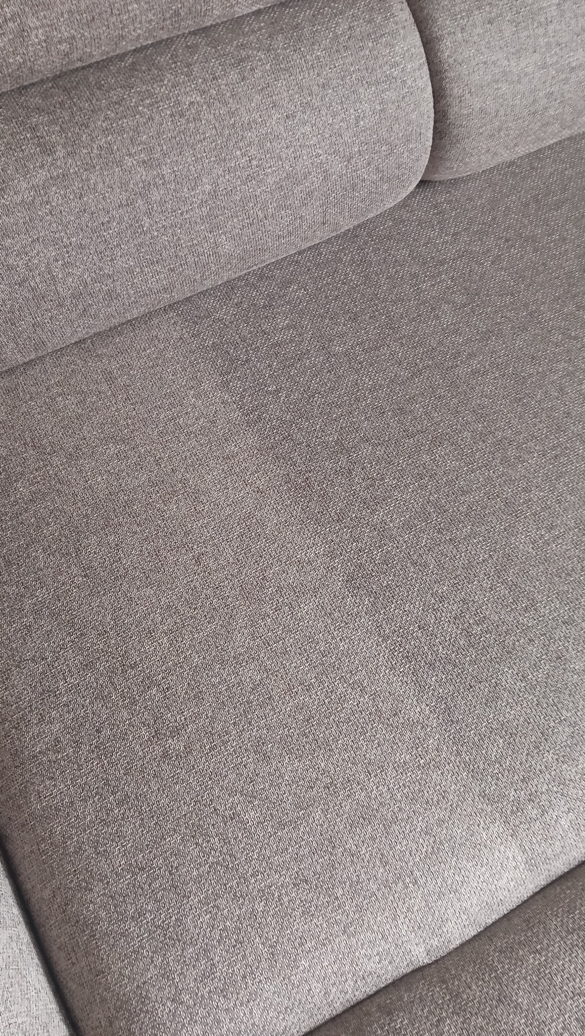 Szczegółowe ujęcie szarej, tekstylnej tapicerki sofy z widoczną fakturą materiału i delikatnym cieniowaniem, ukazujące miękkość i komfort mebla.