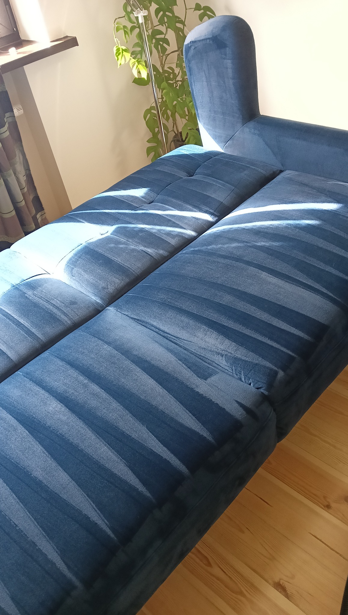 Narożnik z niebieską tapicerką welurową, oświetlony promieniami słońca, stojący na drewnianej podłodze. W tle widoczna roślina doniczkowa i okno.