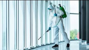 Pracownik w kombinezonie ochronnym, masce i rękawicach dezynfekuje podłogę w jasnym, przeszklonym pomieszczeniu za pomocą opryskiwacza plecakowego.