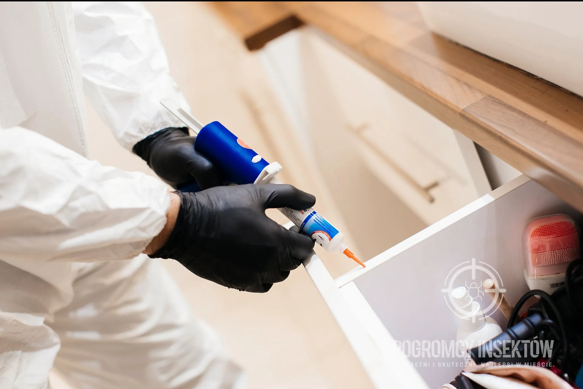 Osoba w kombinezonie ochronnym i czarnych rękawicach aplikuje preparat z tubki do wnętrza otwartej szuflady, widoczna zawartość szuflady i drewniany blat powyżej.