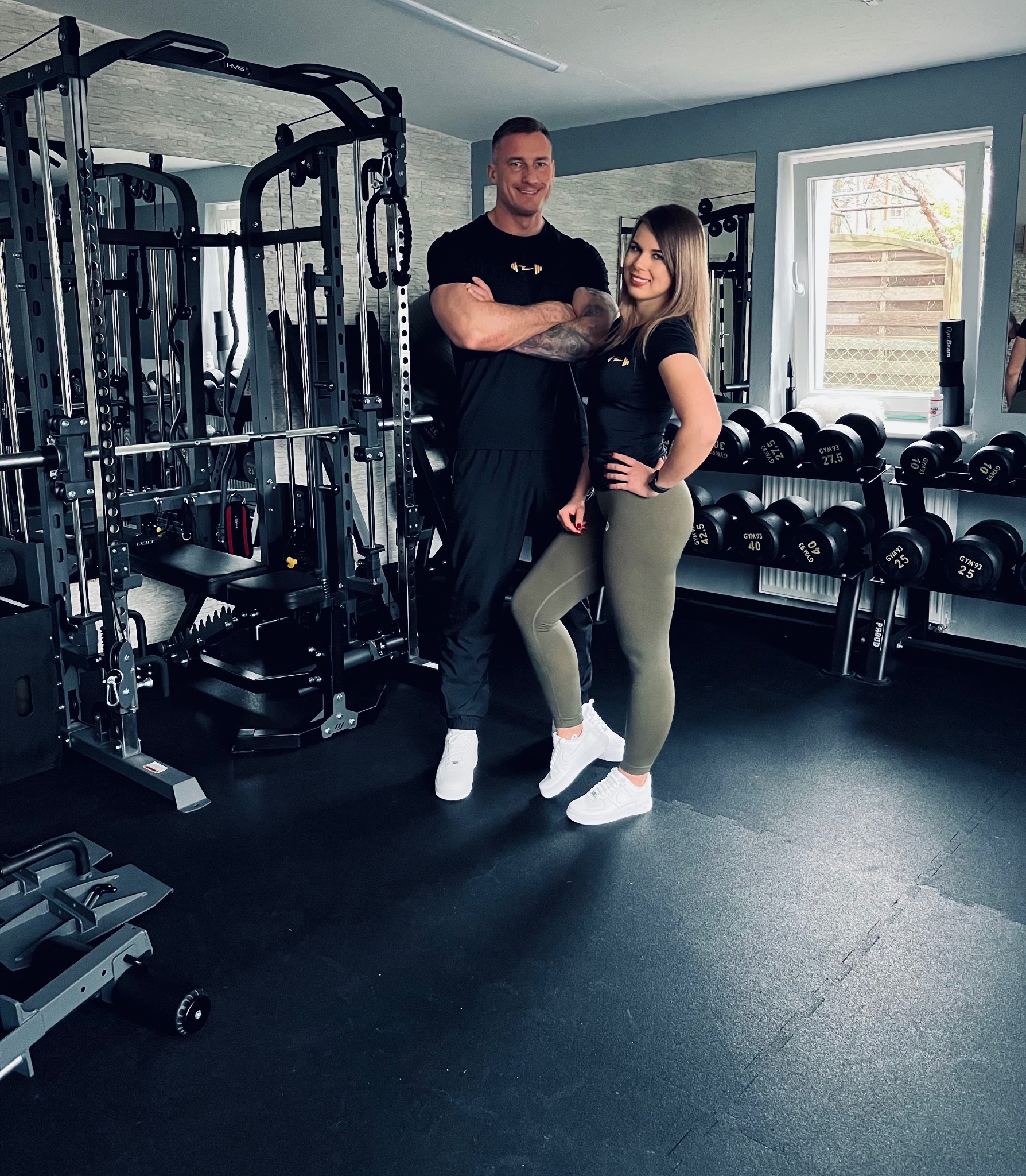 Uśmiechnięty mężczyzna z tatuażami na ramieniu i kobieta w sportowych strojach, pozujący w siłowni na tle sprzętu do ćwiczeń i regału z hantlami.
