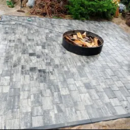 Świeżo ułożona, szara kostka brukowa tworząca duży taras, z czarną obwódką i metalowym paleniskiem wypełnionym drewnem w rogu, otoczona zielenią i stertą gałęzi.