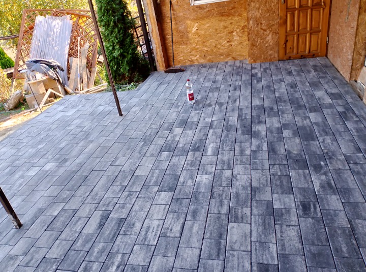 Ułożona szara kostka brukowa na tarasie przylegającym do drewnianego budynku, z butelką wody na środku.