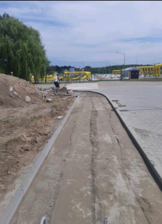 Przygotowanie podłoża pod chodnik z kostki brukowej, widoczne świeżo wylane fundamenty i pracownik z łopatą w tle, na dalszym planie żółta instalacja gazowa.