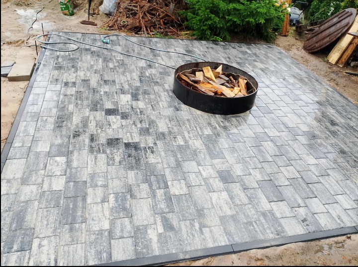 Świeżo ułożona, szara kostka brukowa tworząca duży taras, z czarną obwódką i metalowym paleniskiem wypełnionym drewnem w rogu, otoczona zielenią i stertą gałęzi.