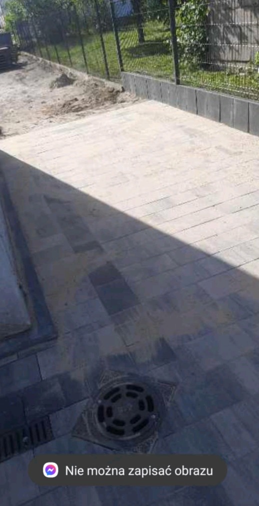 Niedokończona nawierzchnia z kostki brukowej w odcieniach szarości, z widocznym wpustem kanalizacyjnym i piaskiem na powierzchni, obrzeża z betonowych elementów, w tle trawnik i ogrodzenie.