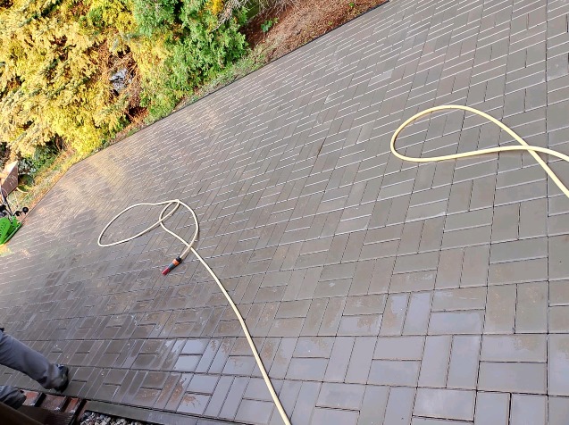 Szare, prostokątne kostki ułożone w jodełkę, mokre od wody z węża ogrodowego, widoczne fragmenty narzędzi i butów osoby wykonującej prace.