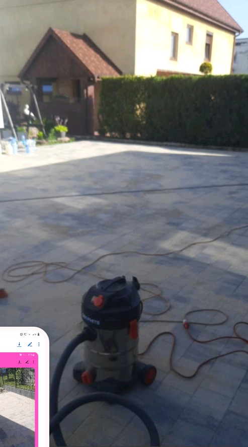 Szary chodnik z kostki brukowej czyszczony na mokro za pomocą odkurzacza przemysłowego Graphite, w tle budynek z brązowym zadaszeniem i zielony żywopłot.