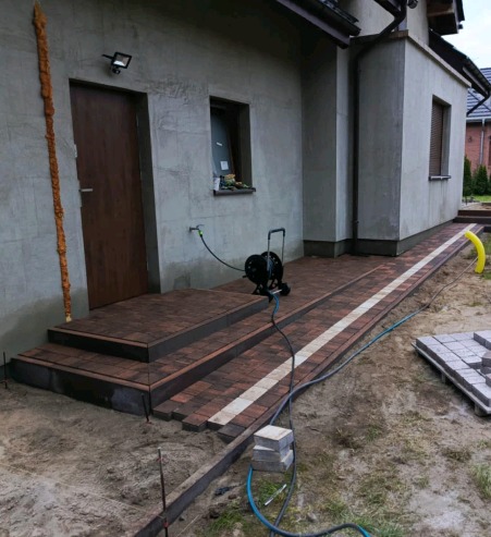 Brązowo-biała kostka brukowa ułożona jako ścieżka i schody przed domem w trakcie prac wykończeniowych, widoczny zwinięty wąż ogrodowy, niezakończone obrzeże i paleta z kostką.