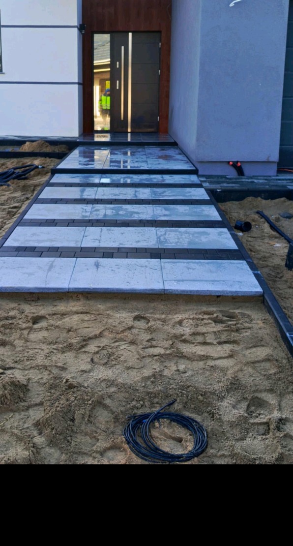 Ścieżka z jasnoszarych płyt i ciemnoszarych kostek prowadząca do nowoczesnego domu, widoczne przygotowane podłoże z piasku i zwój kabla.