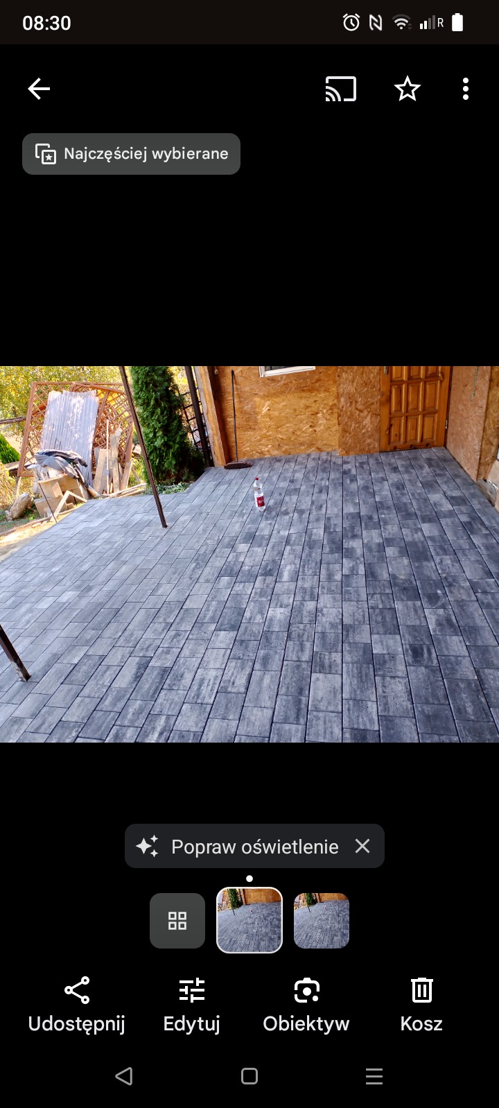 Szara kostka brukowa ułożona przed drewnianym budynkiem, z butelką wody na środku i widoczną drabinką z boku.