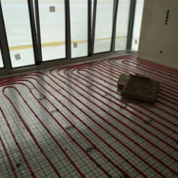 Montaż ogrzewania podłogowego z czerwonych rurek na siatce w pomieszczeniu z dużymi oknami, widoczne deski przy ścianie.