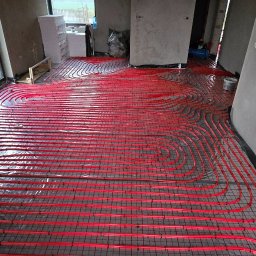 Stawny-Instal Marta Stawna - Czerwone rury ogrzewania podłogowego ułożone na metalowej siatce w surowym wnętrzu, widoczne okno i ściany w stanie deweloperskim.