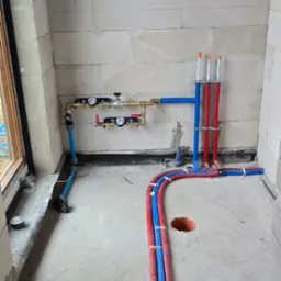 Surowy stan instalacji wodnej i grzewczej w trakcie budowy, widoczne rury PEX w kolorach niebieskim i czerwonym, połączone z rozdzielaczem i przygotowane do podłączenia, ściany z betonu komórkowego
