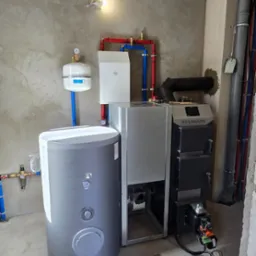 Kocioł na paliwo stałe marki Stalmark, zasobnik ciepłej wody, naczynie przeponowe i instalacja grzewcza w surowym pomieszczeniu z betonową posadzką i ścianami w trakcie wykończenia.