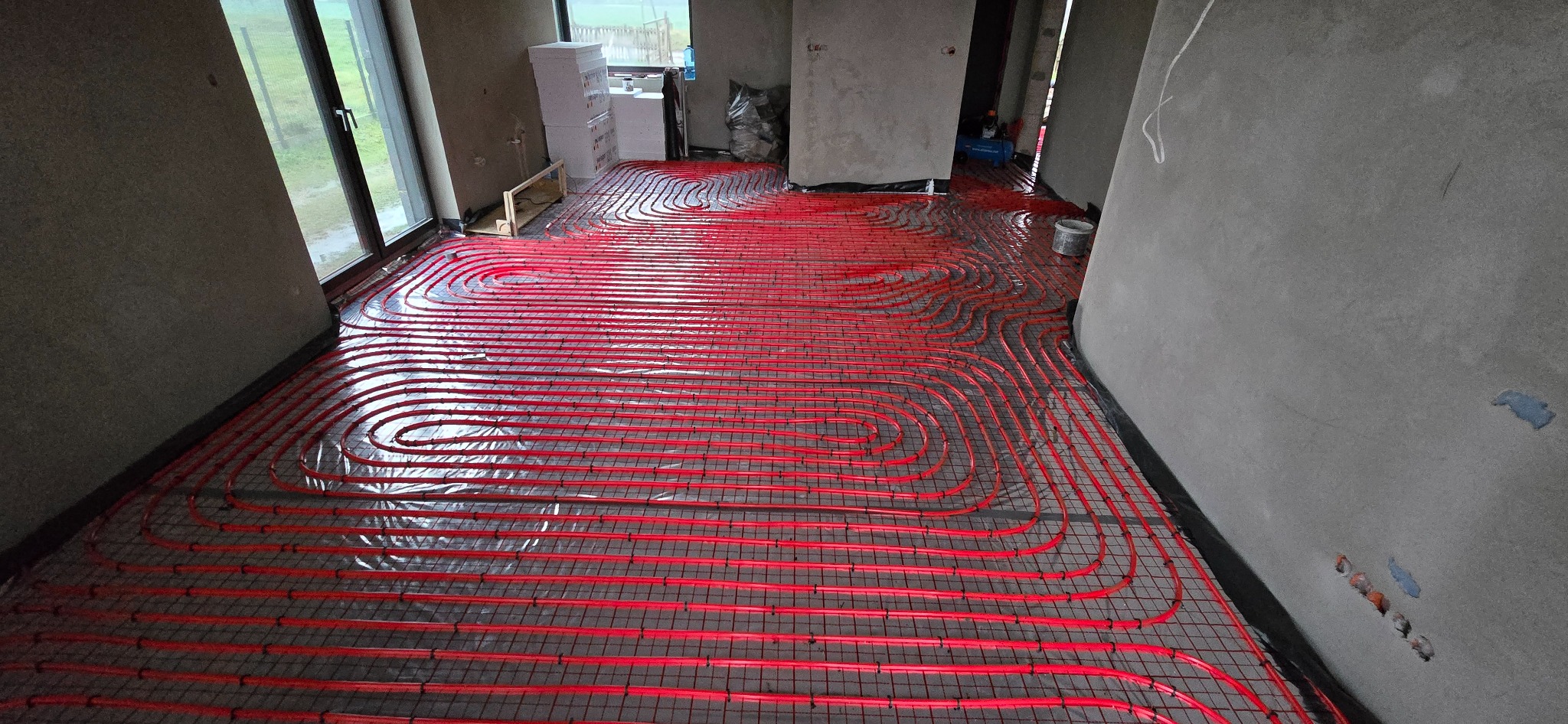 Czerwone rury ogrzewania podłogowego ułożone na metalowej siatce w surowym wnętrzu, widoczne okno i ściany w stanie deweloperskim.