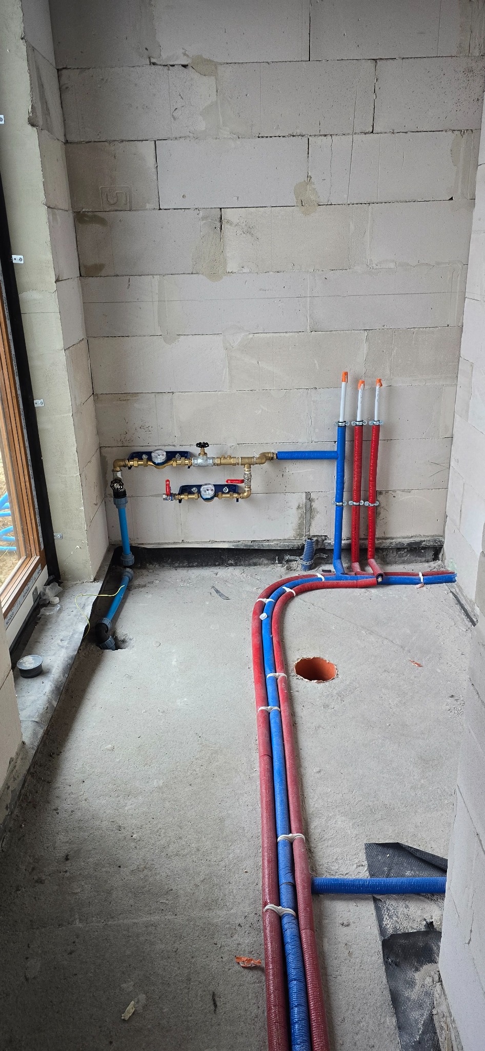 Surowy stan instalacji wodnej i grzewczej w trakcie budowy, widoczne rury PEX w kolorach niebieskim i czerwonym, połączone z rozdzielaczem i przygotowane do podłączenia, ściany z betonu komórkowego