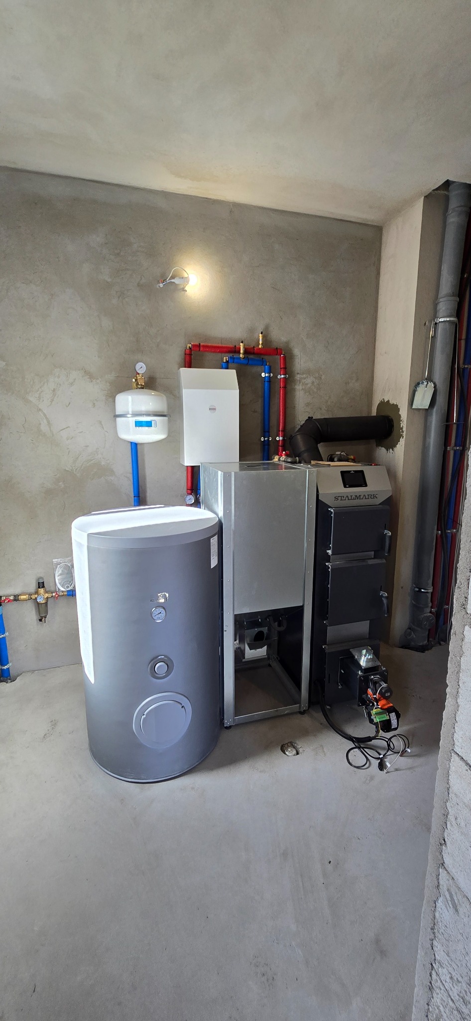 Kocioł na paliwo stałe marki Stalmark, zasobnik ciepłej wody, naczynie przeponowe i instalacja grzewcza w surowym pomieszczeniu z betonową posadzką i ścianami w trakcie wykończenia.