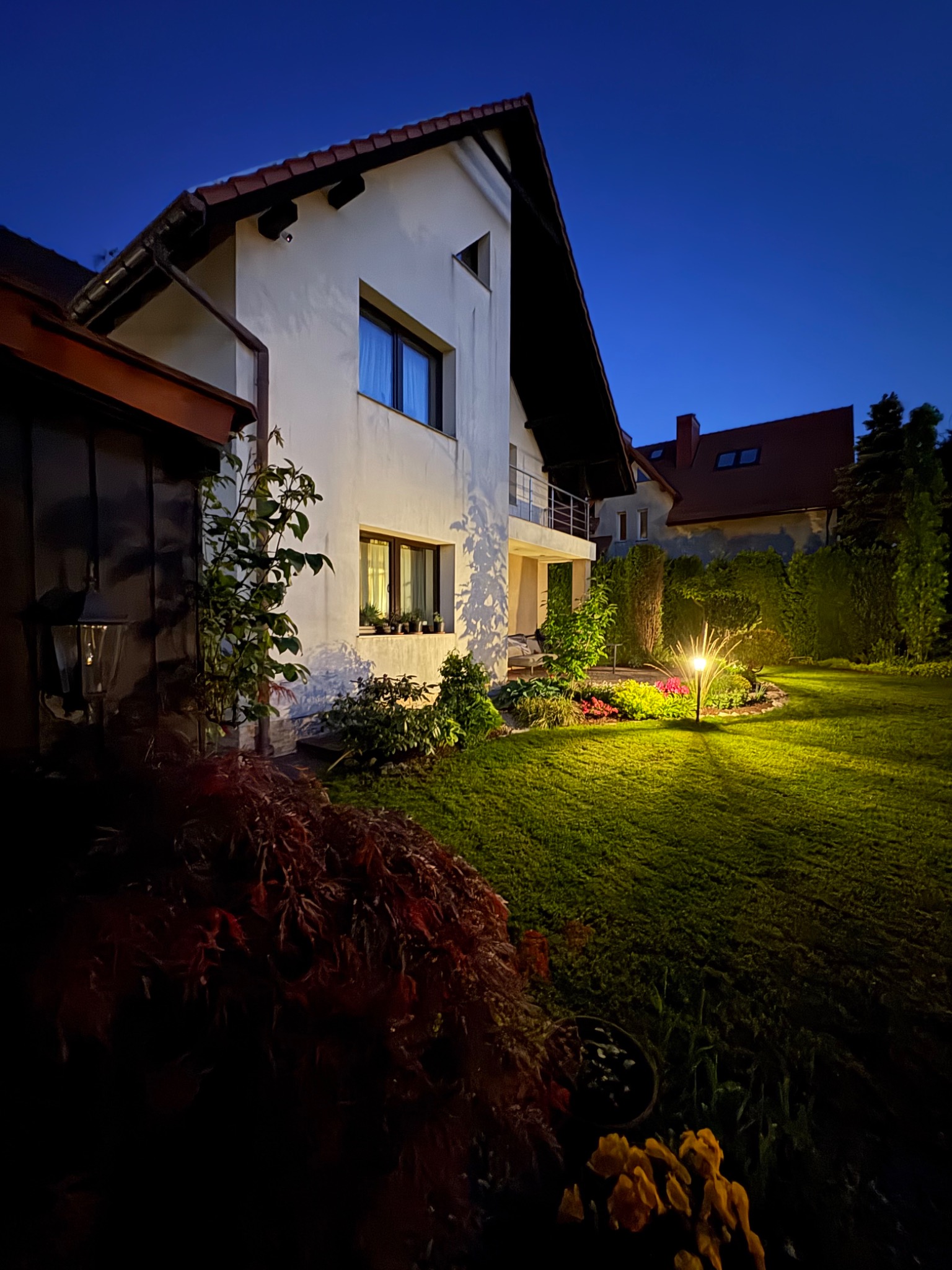 Oświetlony ogród przydomowy o zmierzchu, z lampą ogrodową, rabatami kwiatowymi i zadbanym trawnikiem na tle białego domu z balkonem.