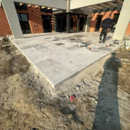 Wykończony taras z jasnoszarej kostki na posesji z nowoczesnym budynkiem z cegły, widoczne narzędzia i cień osoby podczas prac wykończeniowych.