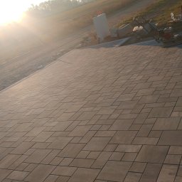 Spec-Kop Patryk Zalewski - Ułożona, szara kostka brukowa na tle łąki w promieniach wschodzącego słońca, widoczna ubijarka wibracyjna i paleta z kostką.