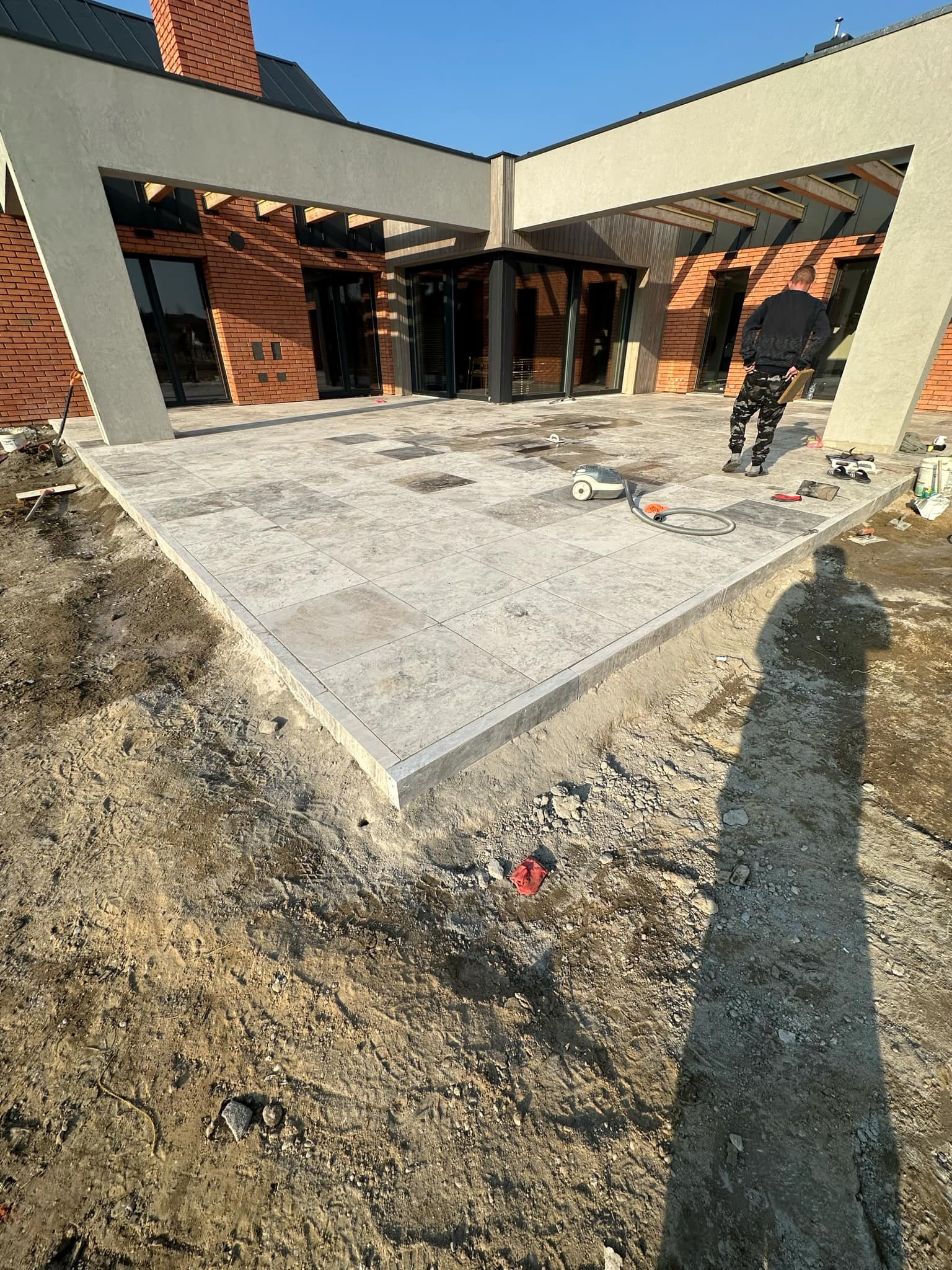 Wykończony taras z jasnoszarej kostki na posesji z nowoczesnym budynkiem z cegły, widoczne narzędzia i cień osoby podczas prac wykończeniowych.