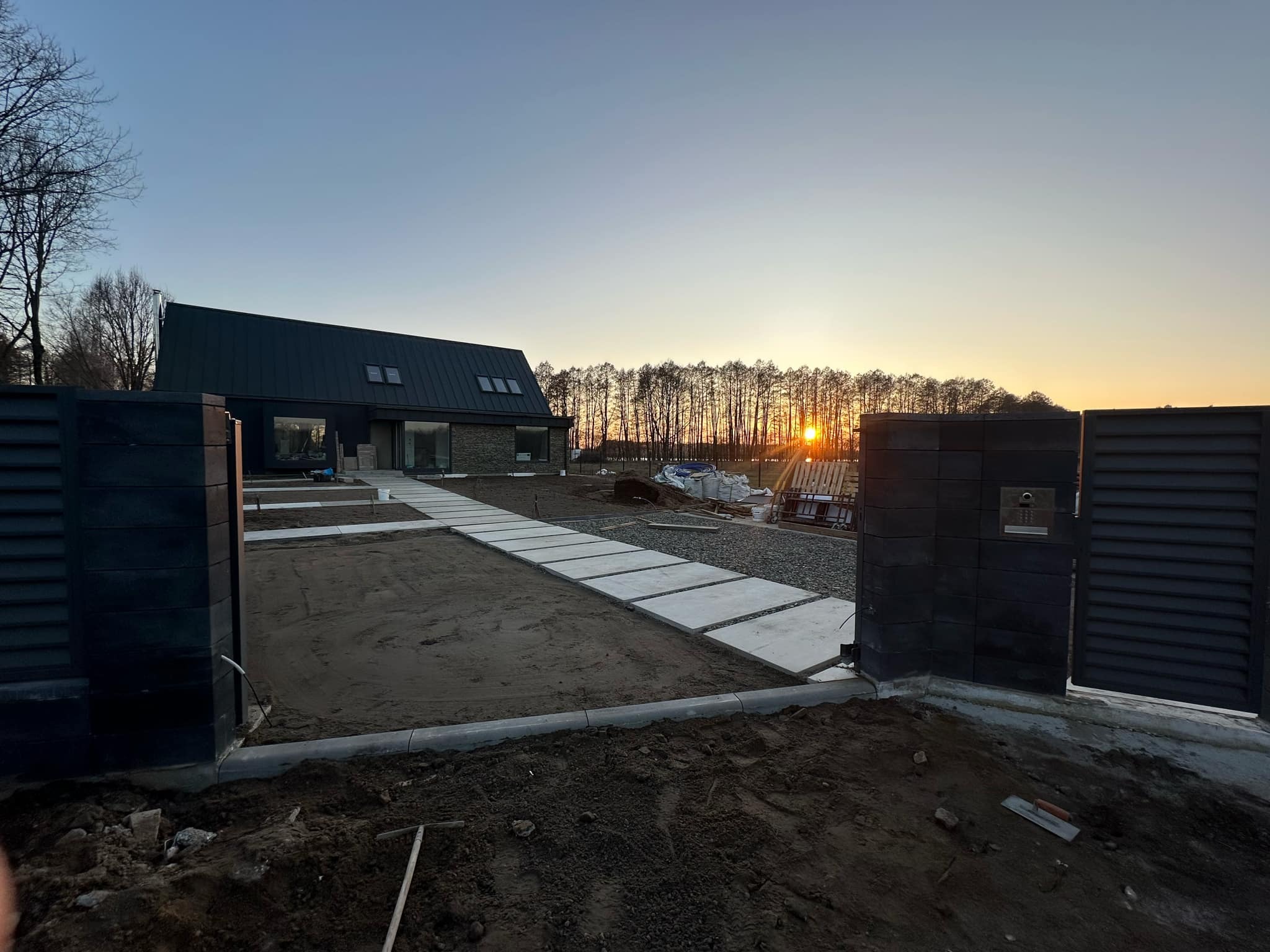 Nowoczesna posesja w trakcie wykańczania: brama wjazdowa z szarej kostki, ścieżka z betonowych płyt prowadząca do domu, widok na zachodzące słońce za drzewami w tle.