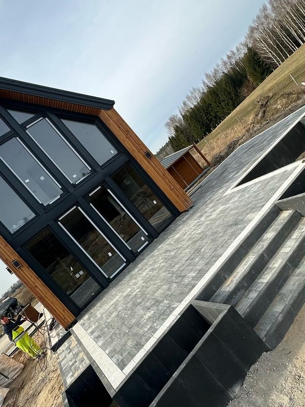 Nowoczesny dom z dużymi oknami i tarasem wyłożonym szarą kostką brukową, widoczne schody z obrzeżami, pracownik w żółtych spodniach i narzędzia w lewym dolnym rogu, w tle drzewa i niebo.