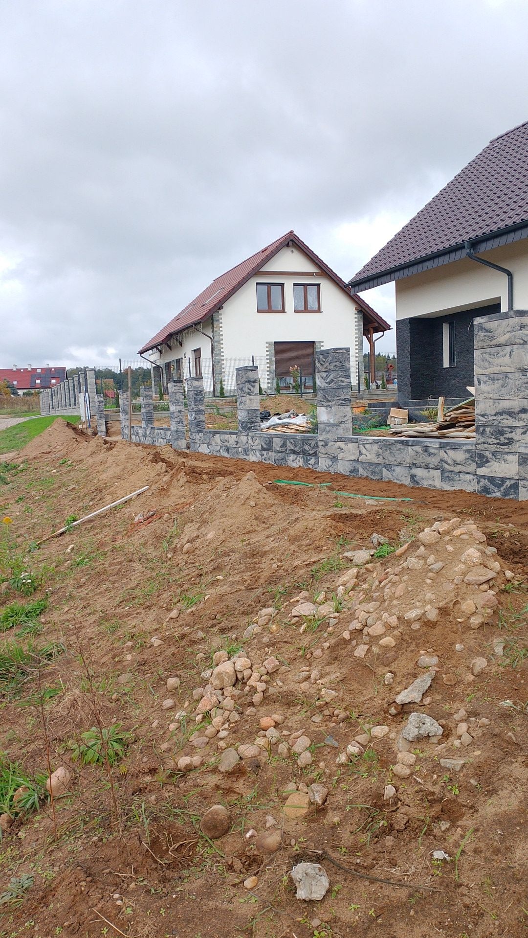 Budowa ogrodzenia z betonowych bloczków imitujących kamień, widoczne dwa domy w tle i wykopana ziemia wzdłuż ogrodzenia.