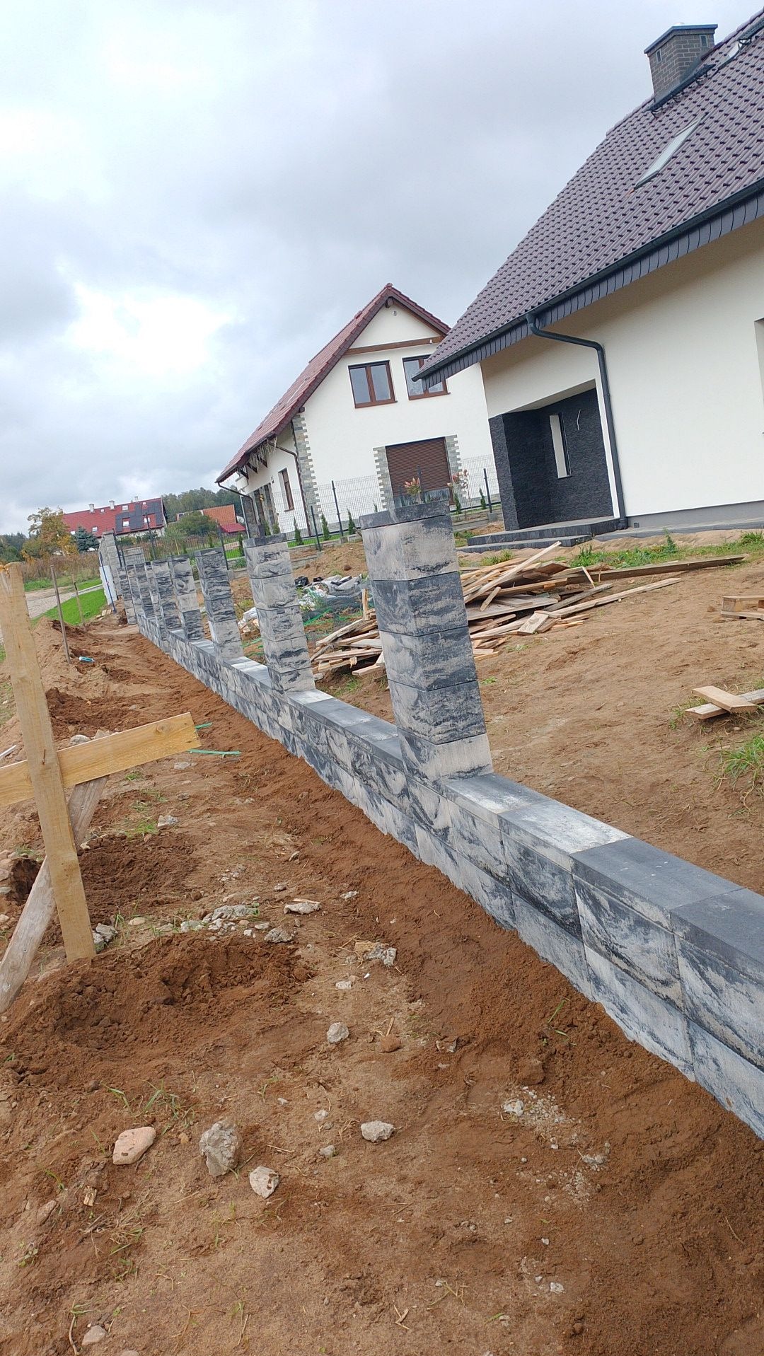Wznoszona linia ogrodzenia z szarej kostki betonowej, widoczne słupki i podmurówka w trakcie montażu, na pierwszym planie ziemia, w tle domy jednorodzinne.