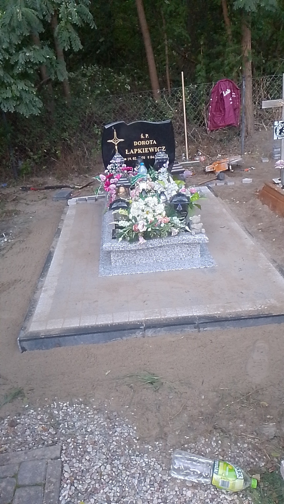 Granitowy nagrobek z kwiatami i zniczami na cmentarzu, otoczony piaskiem i kostką brukową, widoczna butelka po napoju