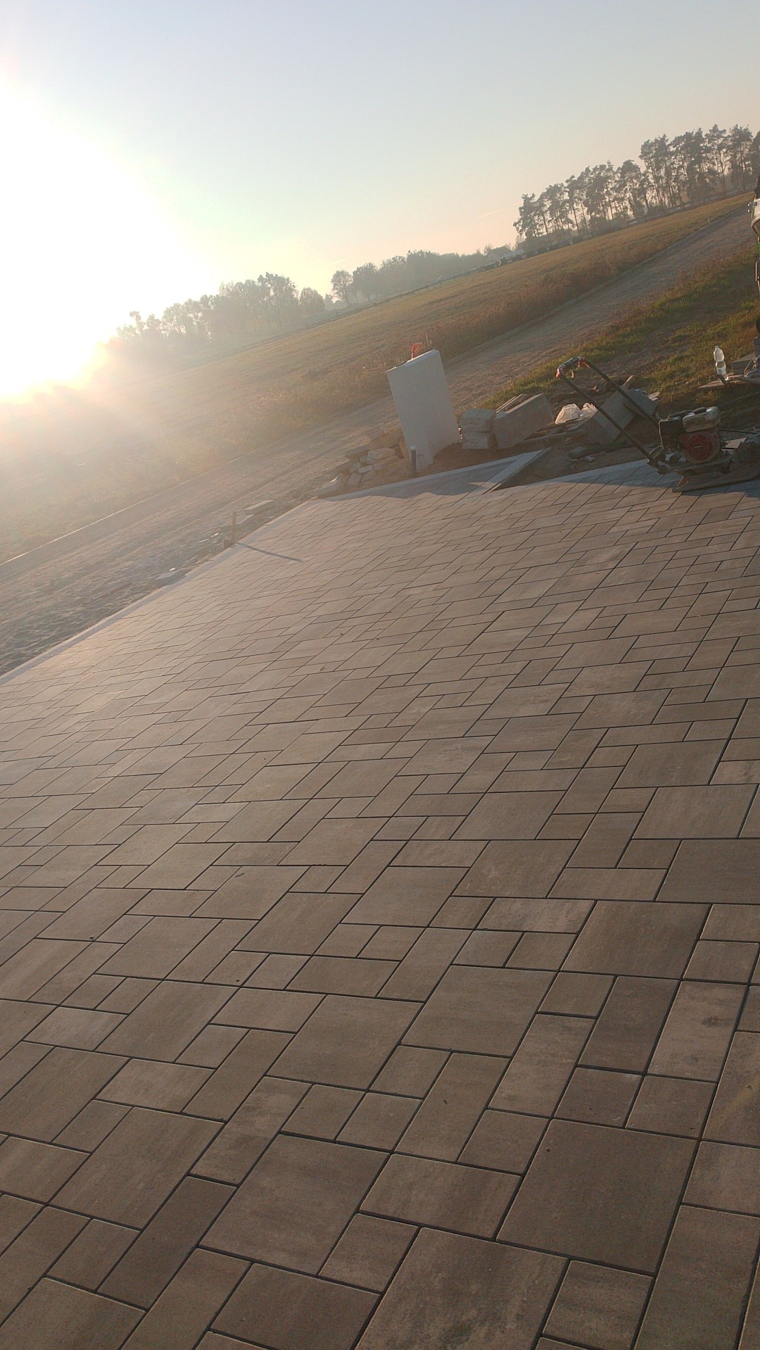 Ułożona, szara kostka brukowa na tle łąki w promieniach wschodzącego słońca, widoczna ubijarka wibracyjna i paleta z kostką.