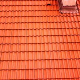 Pomarańczowa dachówka ceramiczna ułożona rzędami, widok z góry, fragment komina z obróbką blacharską w tle.