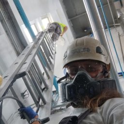 HYDROSERWIS-MALOWANIE, USZCZELNIANIE, CZYSZCZENIE, RENOWACJA DACHÓW, ELEWACJI, KOSTKI - Pracownik w masce ochronnej i kasku, stojący na ziemi, obserwuje drugiego malującego ścianę na wysokości z drabiny w pomieszczeniu przemysłowym z rurami i instalacjami pod sufitem.