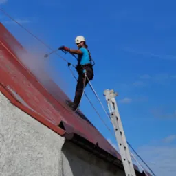 Pracownik w kasku i uprzęży zabezpieczającej czyści czerwoną, metalową dachówkę myjką ciśnieniową, z boku drabina oparta o budynek.