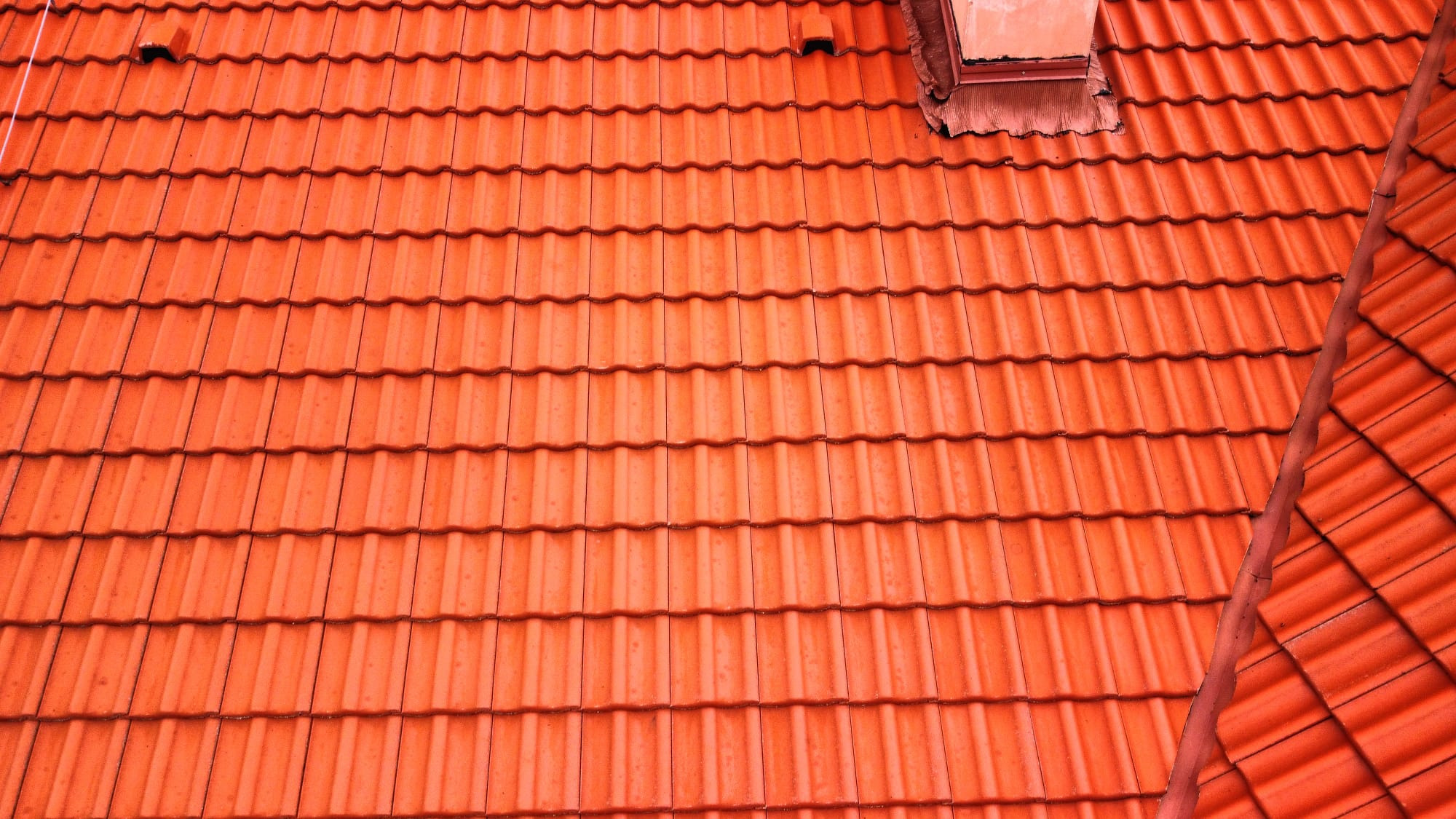 Pomarańczowa dachówka ceramiczna ułożona rzędami, widok z góry, fragment komina z obróbką blacharską w tle.