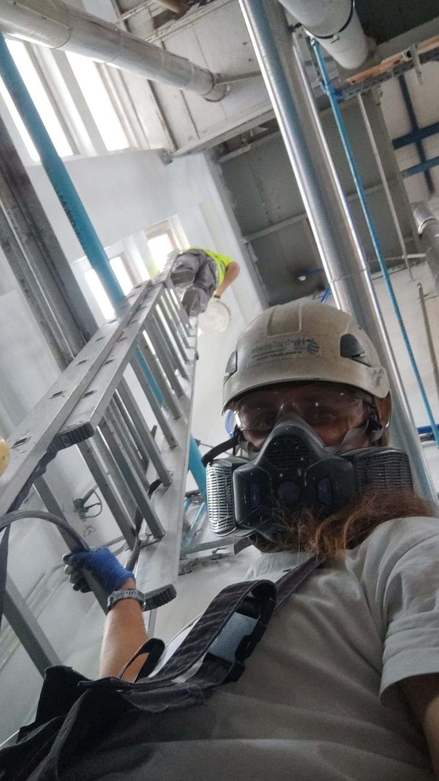 Pracownik w masce ochronnej i kasku, stojący na ziemi, obserwuje drugiego malującego ścianę na wysokości z drabiny w pomieszczeniu przemysłowym z rurami i instalacjami pod sufitem.