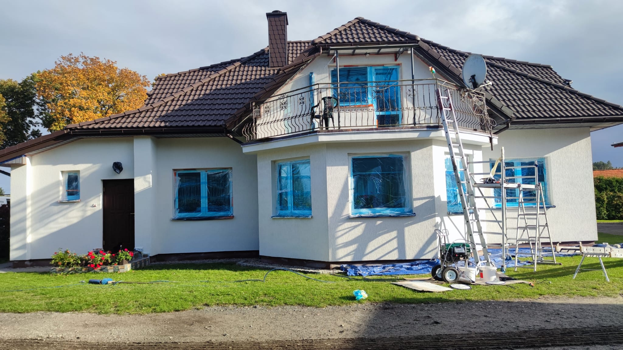 Dom jednorodzinny z balkonem w trakcie przygotowań do malowania elewacji, okna zabezpieczone folią, widoczna drabina, agregat malarski i sprzęt. Zabezpieczony teren wokół budynku.